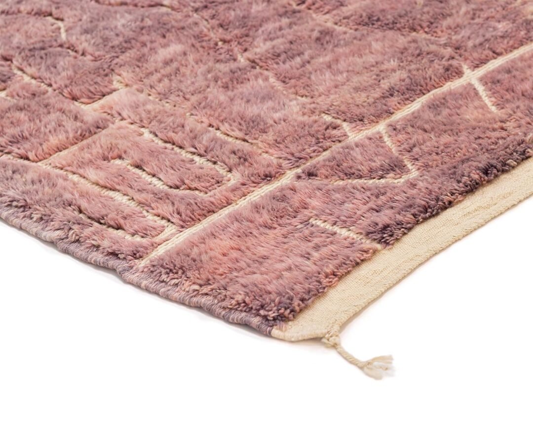 Close-up of a corner of a textured, purple-toned, hand-knotted rug with a geometric pattern and a tassel at the end.