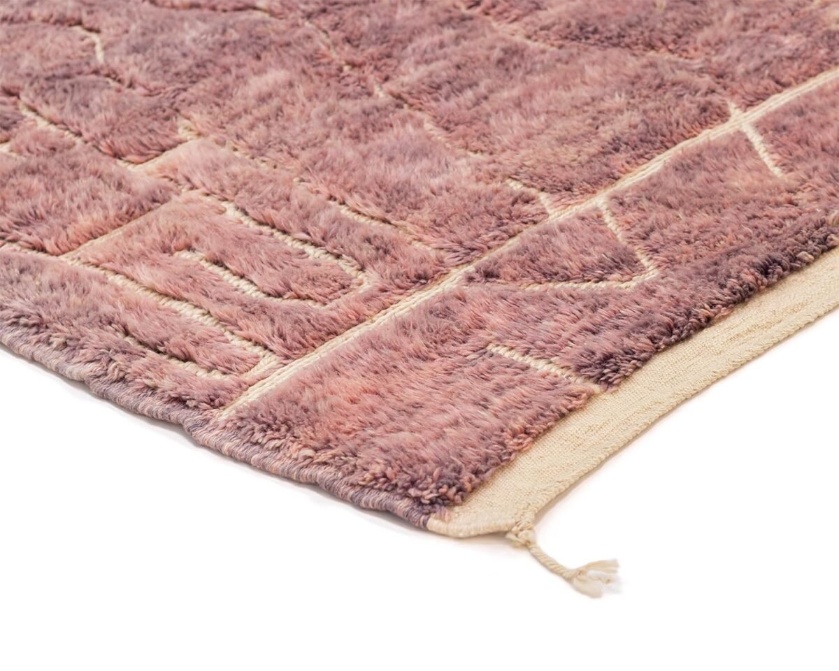 Close-up of a corner of a textured, purple-toned, hand-knotted rug with a geometric pattern and a tassel at the end.