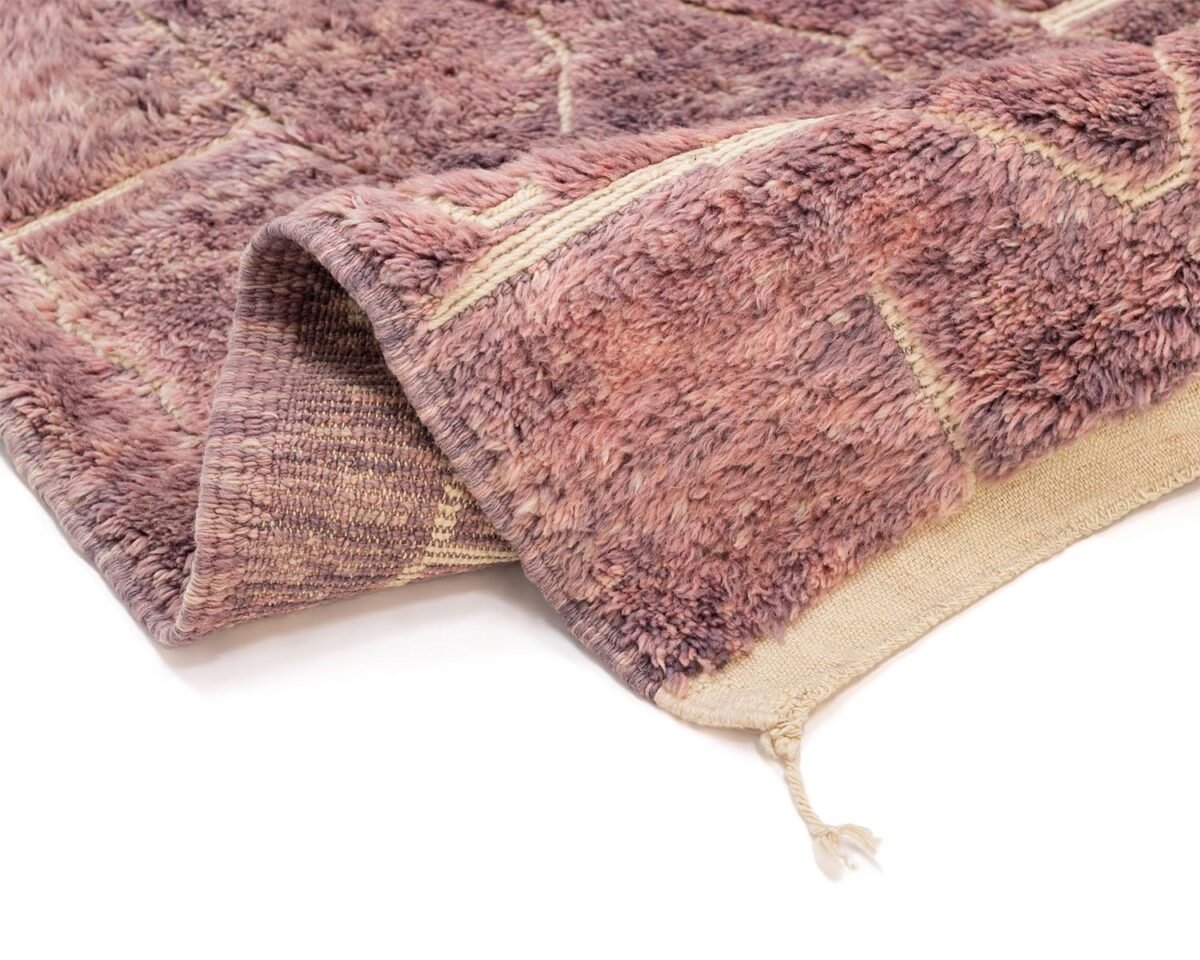 A folded, textured hand-knotted rug with a geometric pattern in shades of pink and beige, featuring a fringe on one corner.