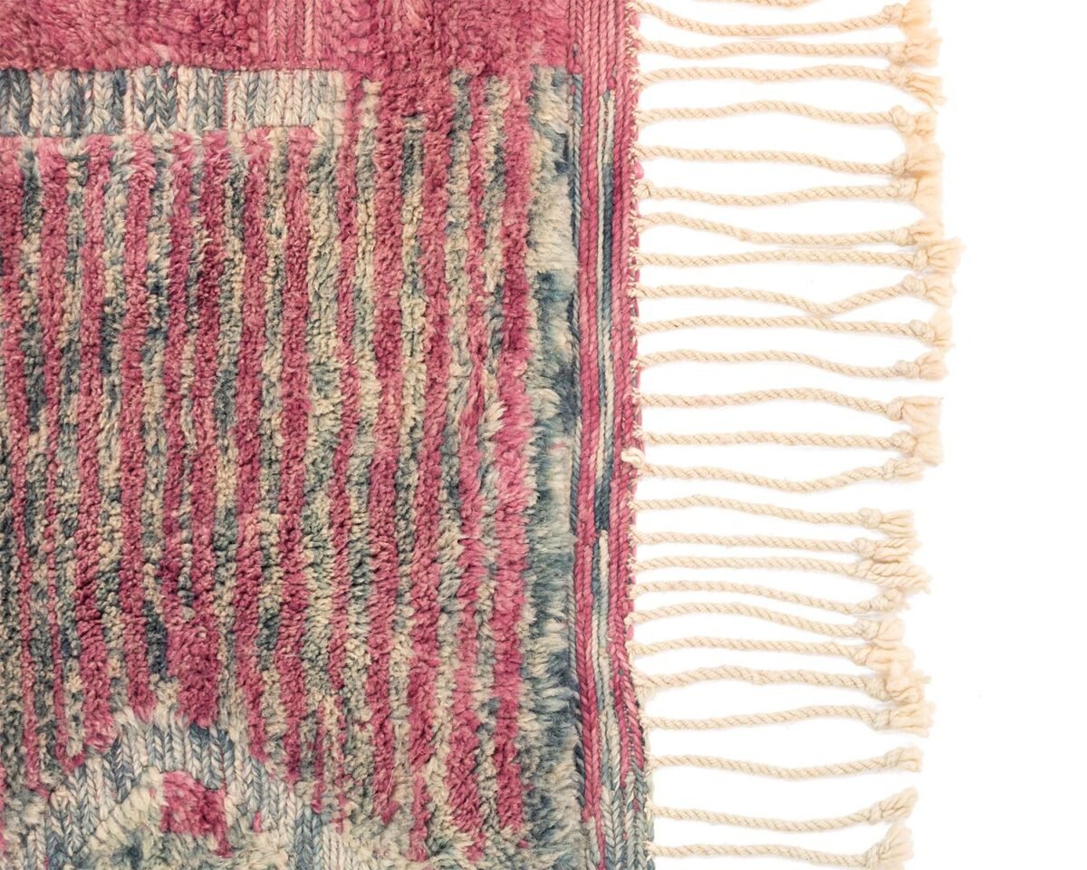 This luxurious Moroccan rug features a textured pattern in pink, blue, and beige. On the right side, cream-colored fringe tassels add an elegant touch.