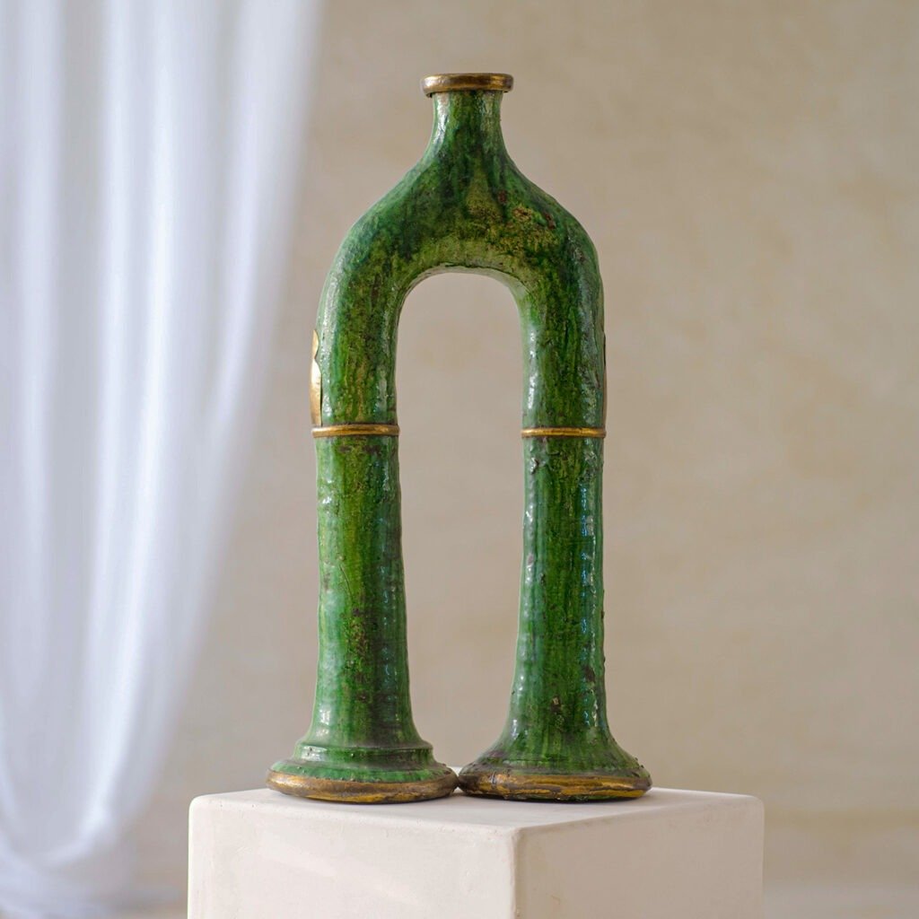 A jade-green, arch-shaped ceramic vase with a textured surface stands elegantly on a white pedestal, featuring a soft-focus white curtain delicately draped in the background.