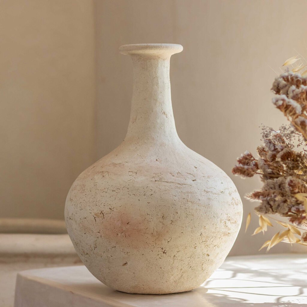 A textured, beige ceramic vase with a narrow neck and round body stands on a light surface, complemented by a bunch of dried flowers, exuding an artisanal charm.