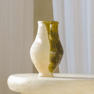 A ceramic vase adorned with a white and olive green glaze rests elegantly on a round, light-toned surface, with soft white curtains serving as the backdrop.