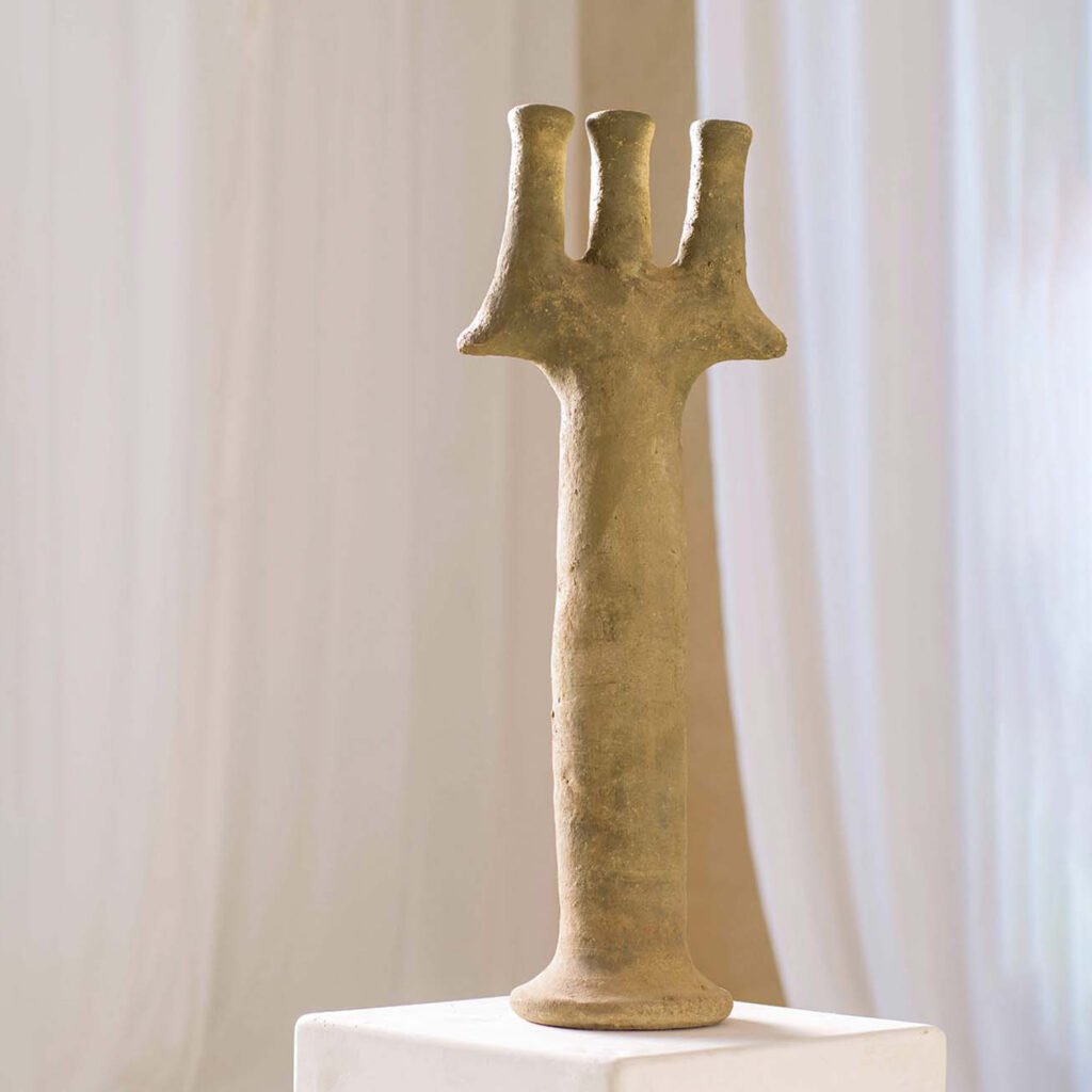 A beige ceramic sculpture resembling a trident stands elegantly on a white pedestal, with billowing white curtains as its backdrop, creating an artistic focal point in the room.