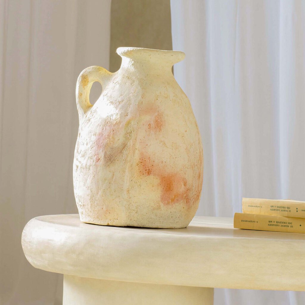 A textured ceramic jug with a handle rests elegantly on a smooth, light-colored table. Nearby, two books peek out, adding an intellectual charm. White curtains gracefully drape in the background.