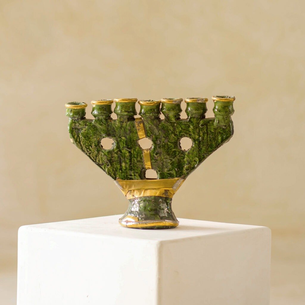 The stunning green and gold ceramic menorah features a textured surface and an asymmetrical design, elegantly showcased on a white pedestal against a neutral background. Its artistic flair brings an exquisite touch to any decor.