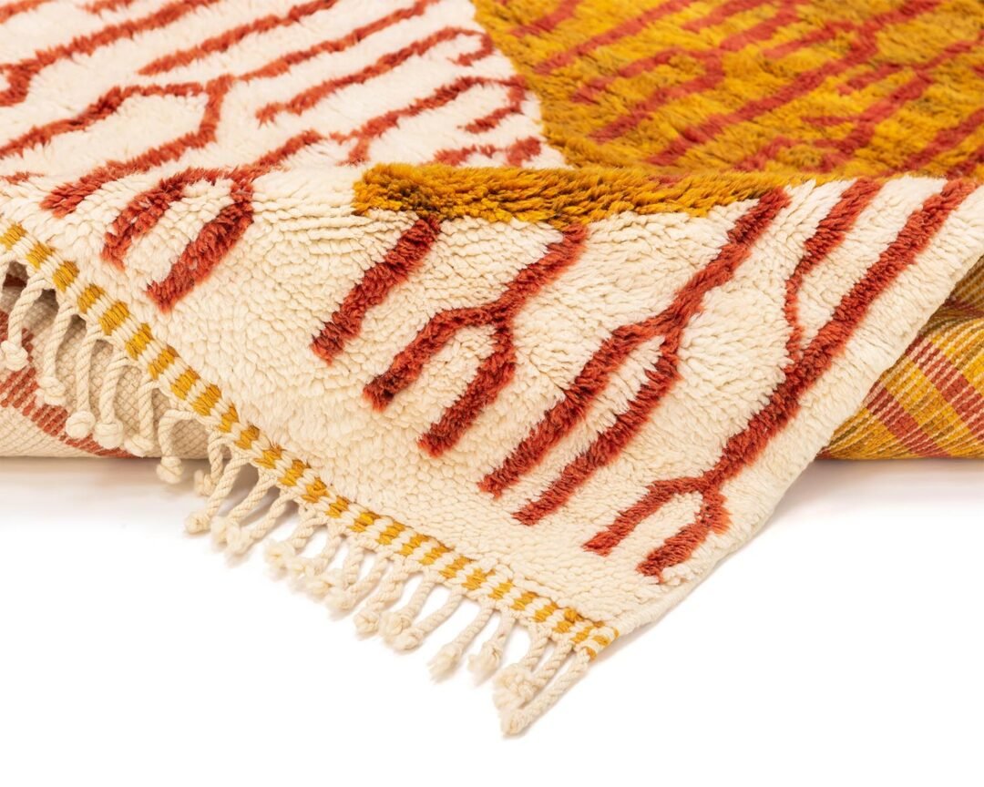 A luxurious, hand-knotted rug with red and orange geometric patterns and fringed edges lies flat on a white surface, epitomizing exquisite handmade home decor.