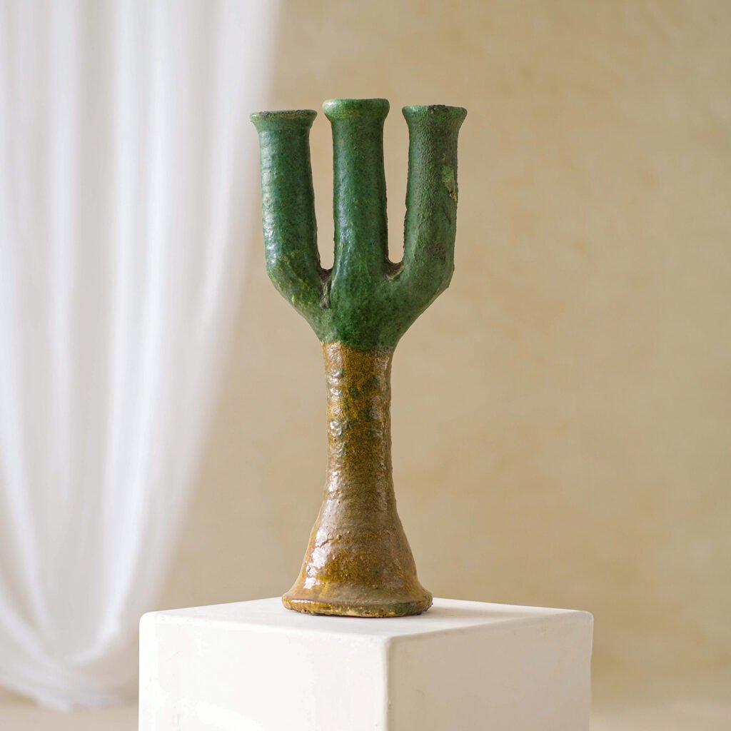 A vibrant, three-pronged ceramic vase in earthy green and brown hues is showcased on a white pedestal, set against a soft backdrop with a gently flowing curtain.