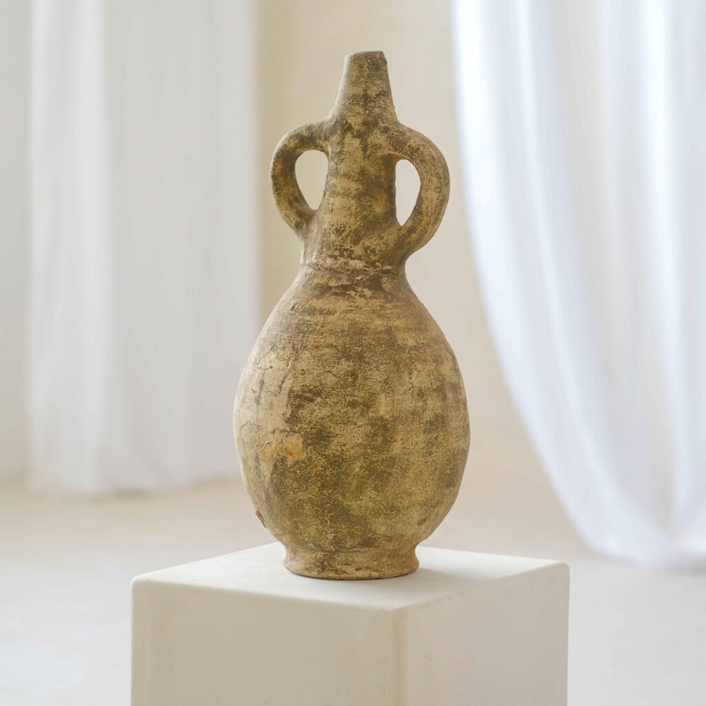 An ancient, weathered ceramic vase with twin handles rests gracefully on a white pedestal against a soft, light backdrop, showcasing its timeless elegance.