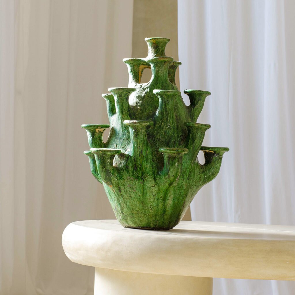 A green, multi-spouted ceramic vase with an organic, layered design graces a smooth, light-colored surface. It stands elegantly against a backdrop of white curtains, adding an artistic touch to the serene setting.