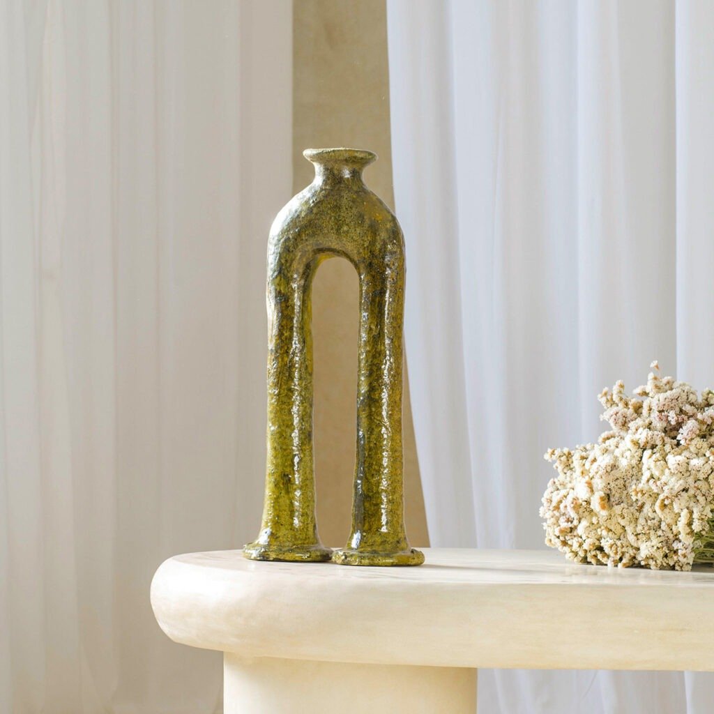 A tall, green ceramic vase with dual legs stands elegantly on a beige table alongside dried flowers, set against the backdrop of a white curtain. The scene exudes a serene and sophisticated charm, enhancing any rooms decor.