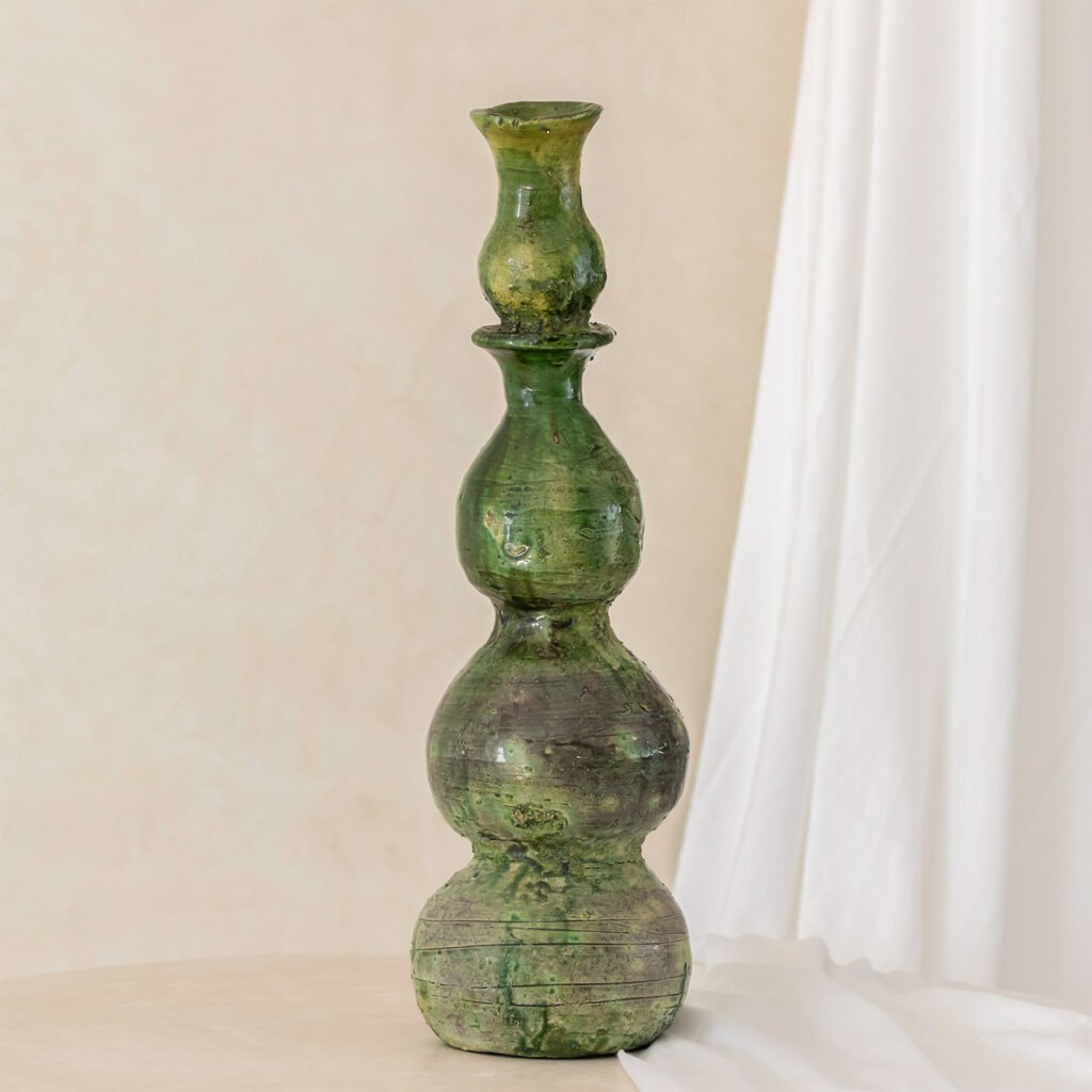 An exquisite antique green glass vase with a multi-tiered, bulbous design stands elegantly against a neutral backdrop enhanced by soft white drapery.
