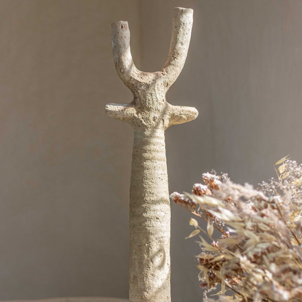 A textured, beige, Y-shaped ceramic sculpture stands upright alongside a cluster of dried flowers on a serene, minimalist background.