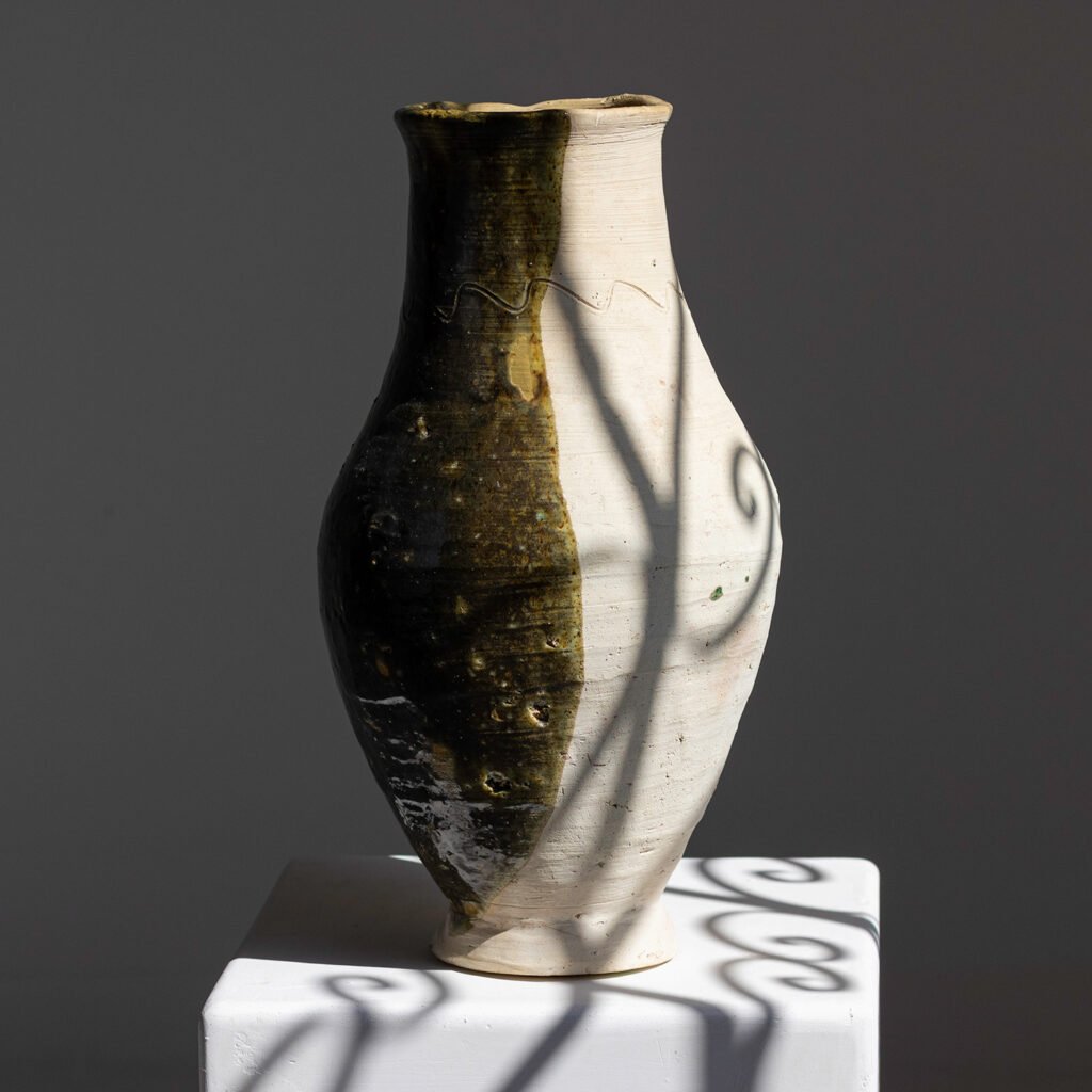 A two-toned ceramic vase sits elegantly on a white pedestal, casting intricate shadow patterns that captivate the eye.
