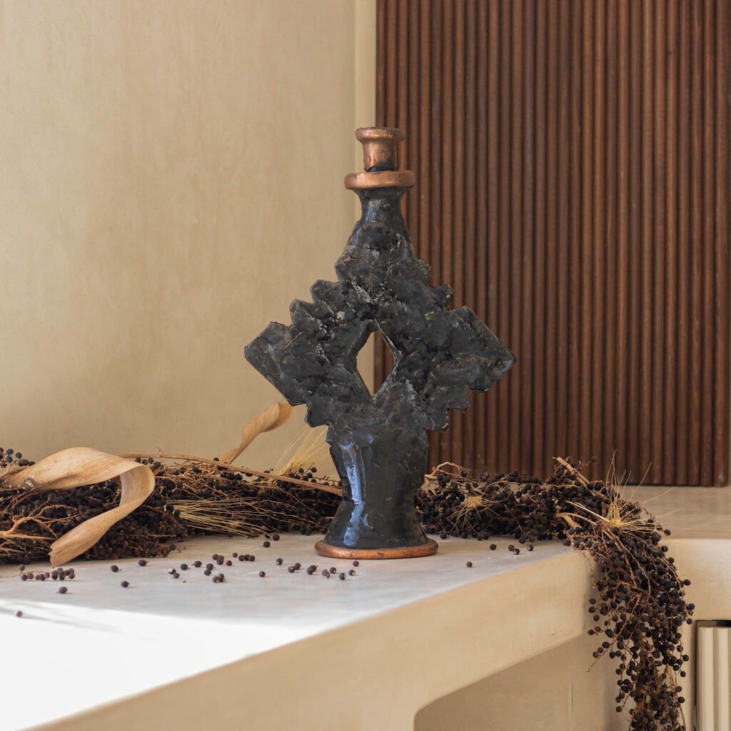 A geometric black sculpture with a textured surface, resembling a bottle, sits on a light-colored surface. Its adorned by dried plants and complemented by wood paneling.