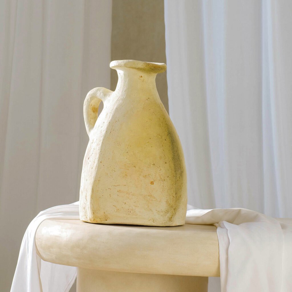 A large, cream-colored ceramic jug reminiscent of Tamegroute pottery with a handle sits on a round beige table, draped with a white cloth, against a white curtain backdrop.