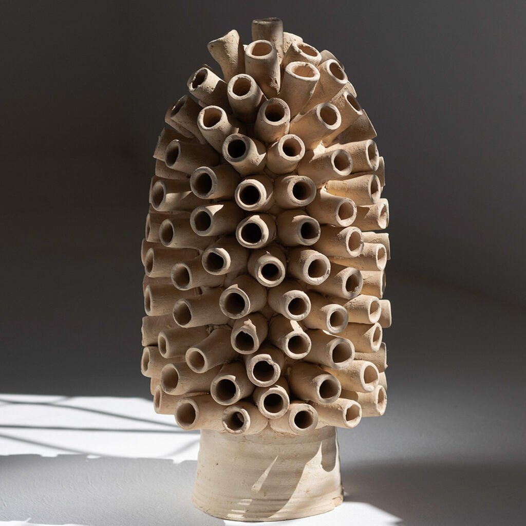 An unglazed Tamegroute pottery sculpture, resembling a cluster of tubes arranged vertically, forms a rounded cone shape, beautifully lit from the side.