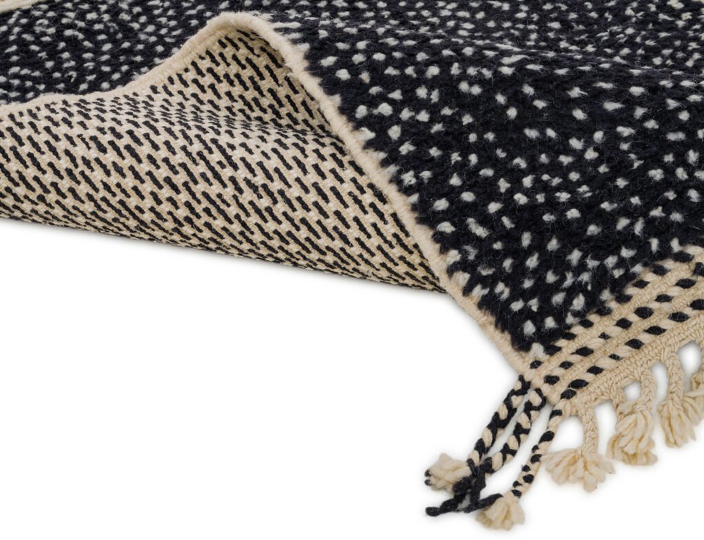 A black and white woven runner rug features a speckled pattern and fringe tassels along the edge, partially folded to reveal its intricate texture.