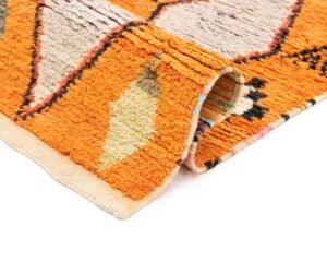 Close-up of an orange runner rug corner with geometric patterns in beige and gray. The fabric is slightly folded, revealing its textured surface and a colorful edge.