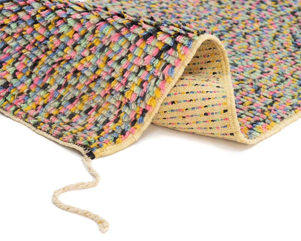 A vibrant woven rug features a folded corner, highlighting its rich texture and multicolored threads. From the edge, a loose yarn strand delicately extends, adding to the intricate detail.