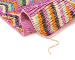 A colorful runner rug with a folded corner, featuring zigzag patterns in pink, orange, green, and black on a white background. A loose thread is visible at the edge.