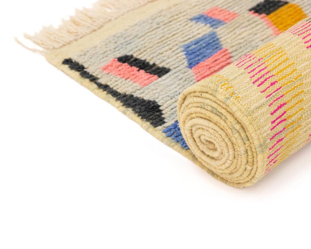 A stylish runner rug with a geometric pattern featuring blue, pink, black, and yellow shapes on a beige background. The rug is partially unrolled, displaying its fringed edges.