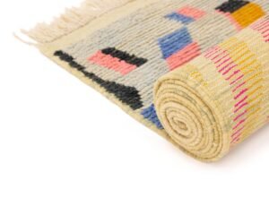 A stylish runner rug with a geometric pattern featuring blue, pink, black, and yellow shapes on a beige background. The rug is partially unrolled, displaying its fringed edges.