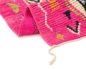 Pink woven runner rug with colorful abstract patterns, featuring a folded corner and a loose string, set against a white background.