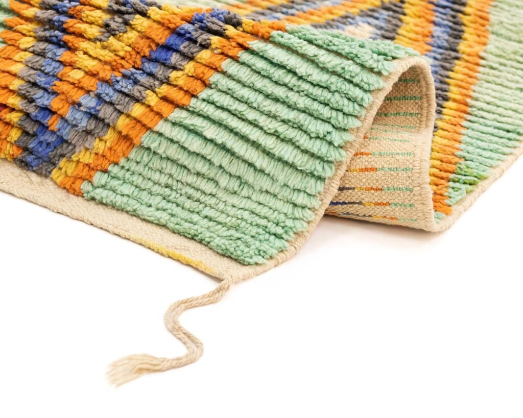 Close-up of a folded, textured woolen runner rug with a geometric pattern in teal, orange, yellow, and blue hues, featuring a loose thread at the bottom corner.