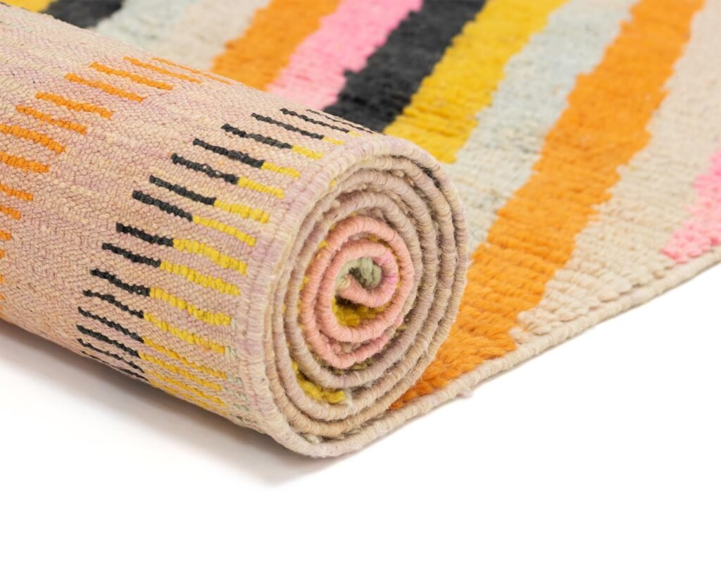 A rolled-up rug with vibrant stripes in orange, pink, black, and yellow lies on a white surface, offering a lively burst of color.
