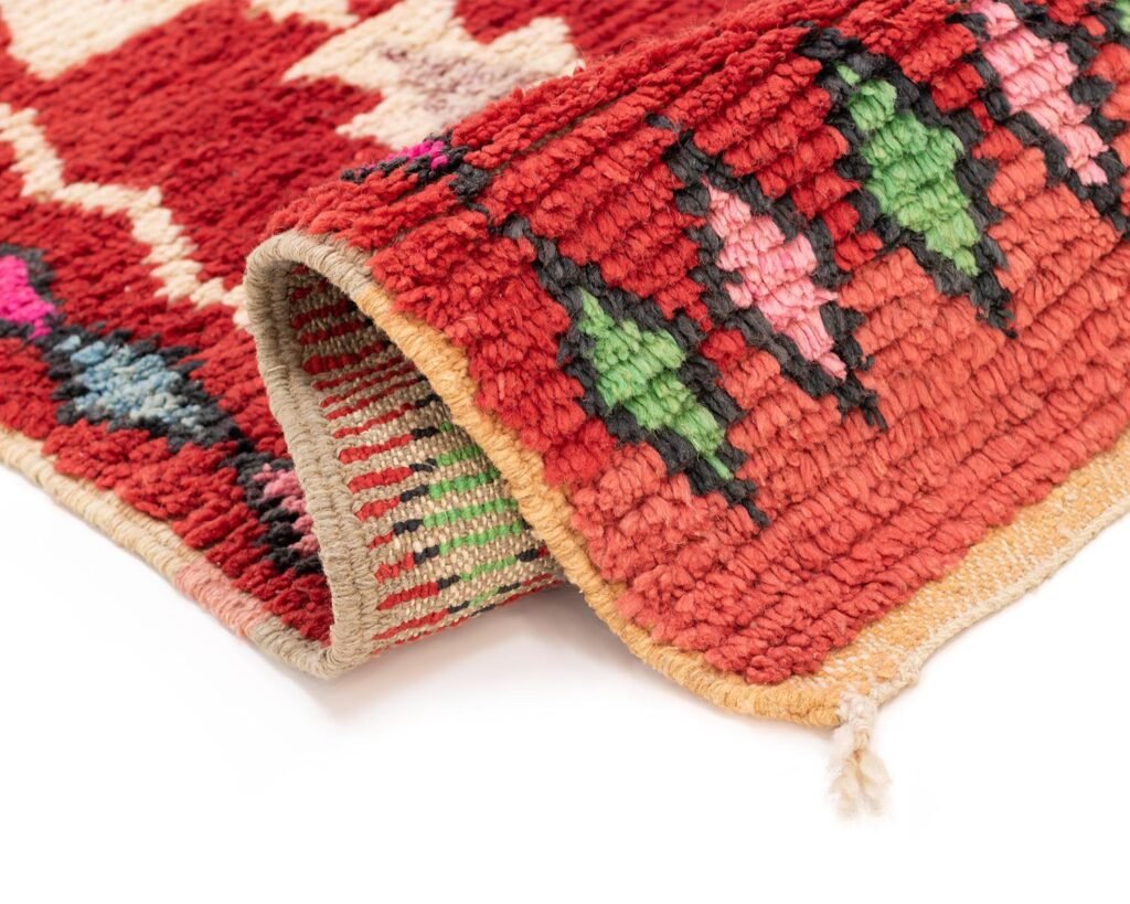 A colorful, textured runner rug corner showcases red, green, black, and pink geometric patterns. The edge is slightly folded to reveal the intricate underside.