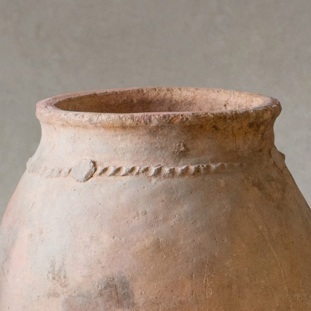 Close-up of the upper section of a weathered clay pot with a visible decorative band around its neck, reminiscent of vintage Berber jars, set against a plain background.