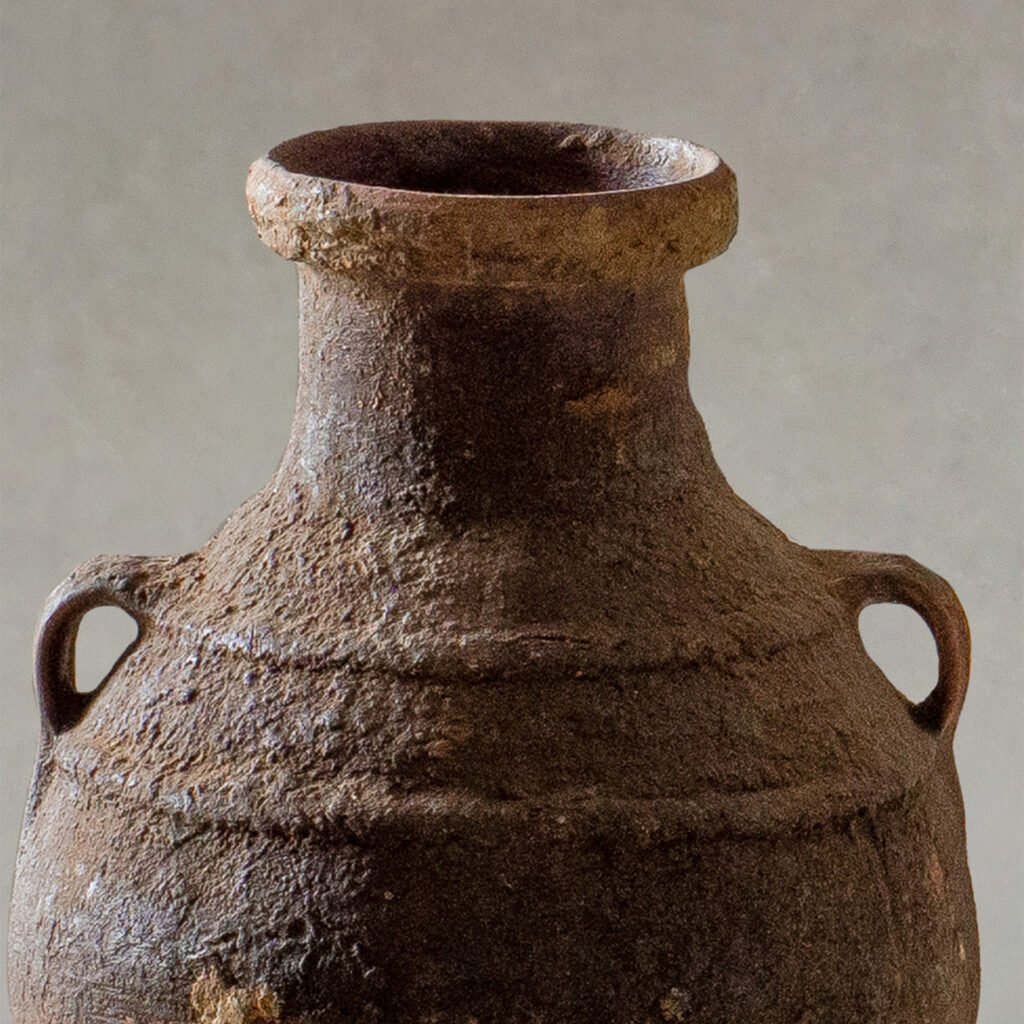 A close-up of an ancient, textured pottery vessel resembles a vintage Berber jar, with two small handles near the top, set against a neutral background.