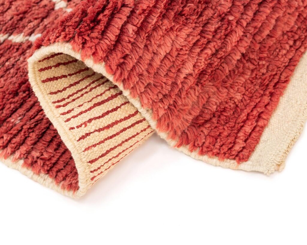 A close-up view of a luxurious red textured rug features a cream underside accented with red stripes. The neatly folded area reveals the cream edge, highlighting the rugs intricate detail and artistry.