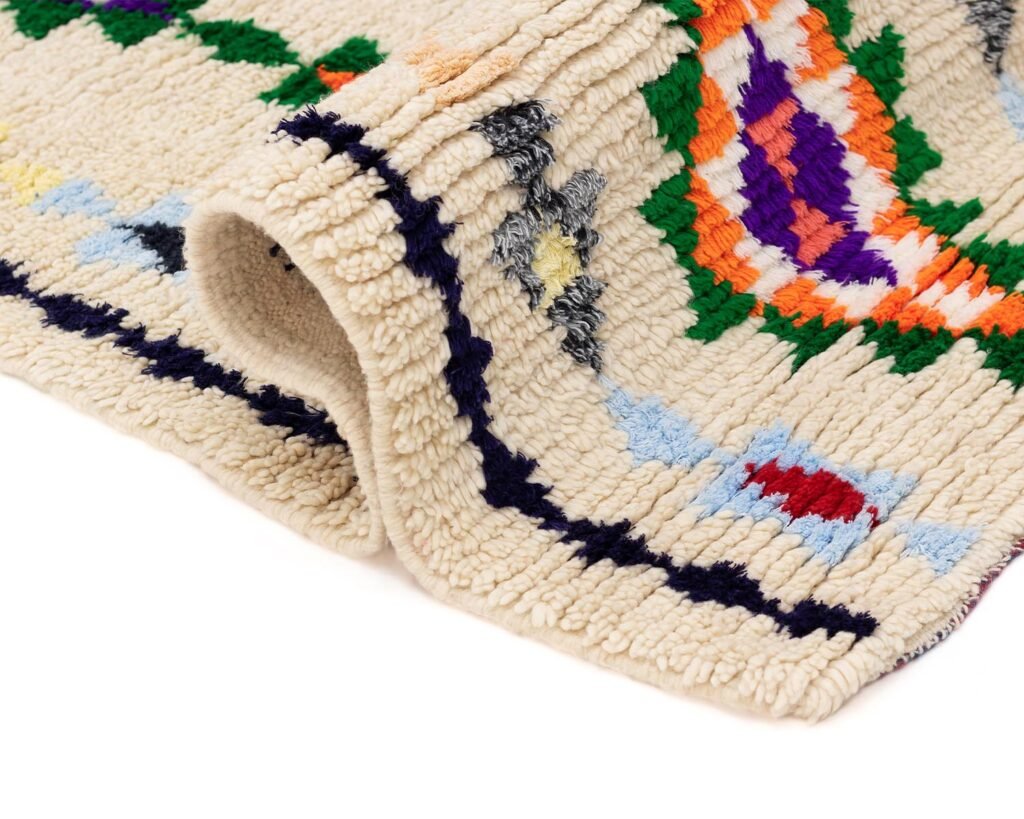 A vibrant woven textile displays abstract patterns in orange, purple, green, blue, and red against a beige background. The folded edge reveals the fabric’s thickness, highlighting its intricate design and craftsmanship.