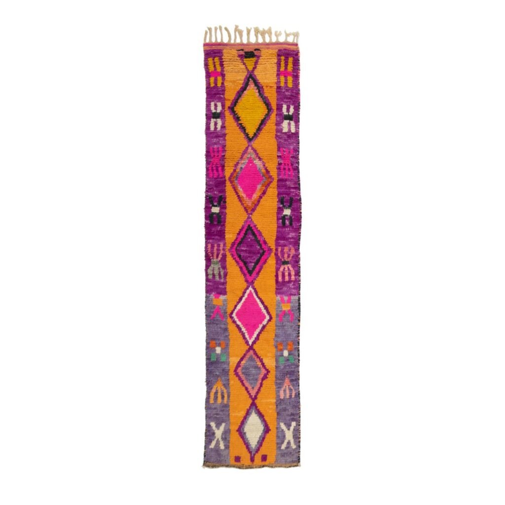 A vibrant, narrow rectangular rug adorned with geometric patterns and fringe on the short edges. It showcases a stunning blend of purple, orange, pink, and gray hues, adding a touch of artistic flair to any room.