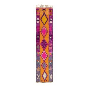 A vibrant, narrow rectangular rug adorned with geometric patterns and fringe on the short edges. It showcases a stunning blend of purple, orange, pink, and gray hues, adding a touch of artistic flair to any room.
