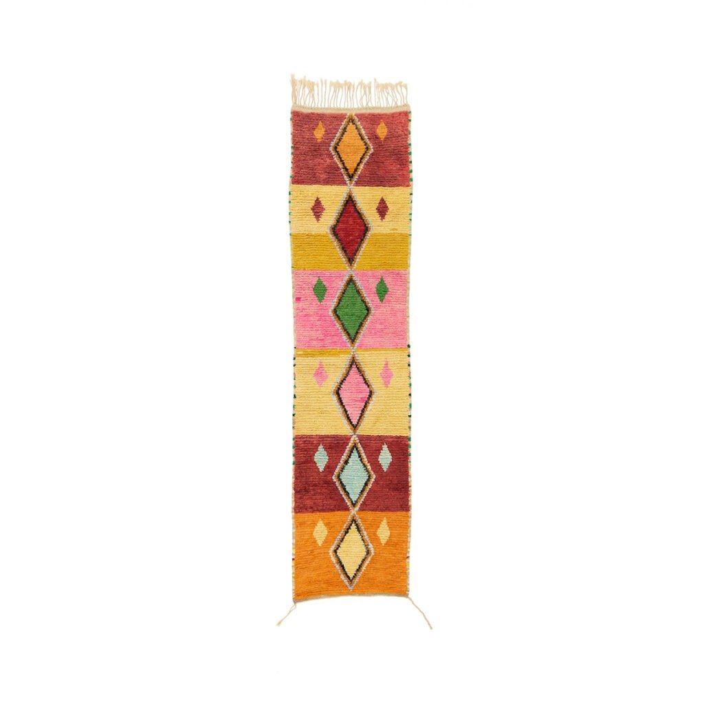 This vibrant runner rug features colorful geometric patterns with diamond shapes and fringe, showcasing shades of red, pink, yellow, green, and orange against a clean white background.