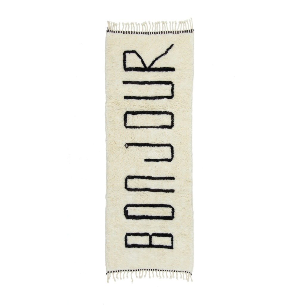 A white rectangular runner rug featuring the word BONJOUR in bold black letters. The edges are elegantly fringed.