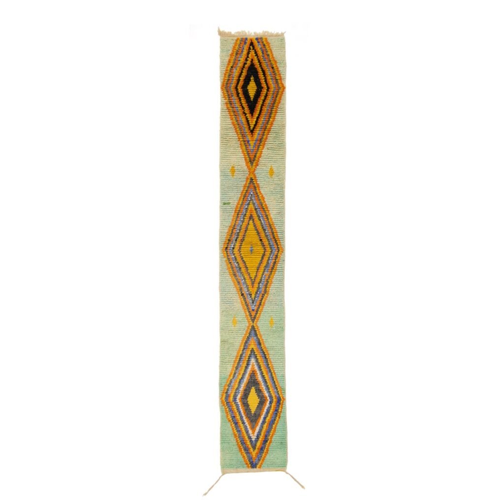 A long, narrow runner rug featuring a series of large, geometric diamond patterns in yellow, blue, and black on a light green background.