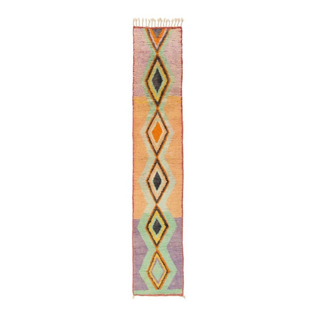 A colorful, narrow runner rug with a geometric diamond pattern in orange, green, yellow, and black hues, featuring fringed edges.