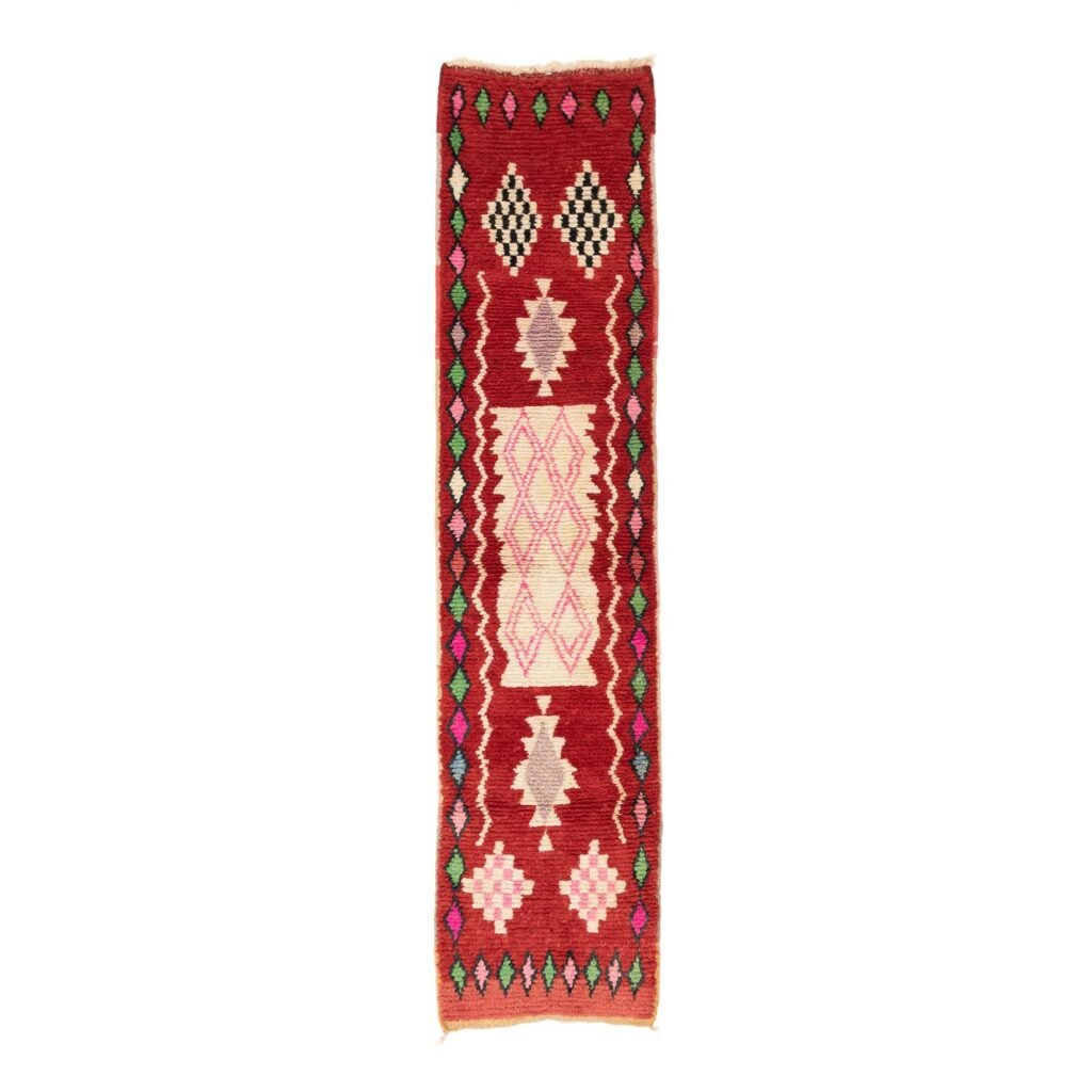 This rectangular runner rug showcases a geometric pattern with diamonds and zigzags in red, pink, black, white, and green on a rich red background.