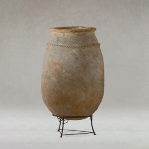 A tall, artisanal clay pot with a textured surface stands elegantly on a metal frame against a neutral backdrop.