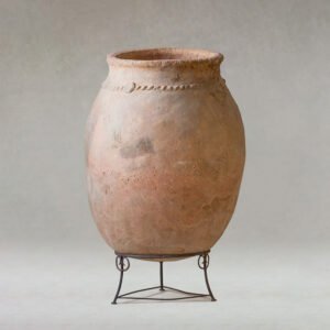 A tall, round, weathered terracotta vase with a slightly textured surface rests elegantly on a simple metal stand against a neutral backdrop. The earthy tones of the vase blend harmoniously into any space.