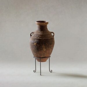 An ancient clay amphora with a narrow neck and two small handles rests gracefully on a simple metal stand, set against a neutral background. Its timeless design evokes an era where such vessels were essential for storing precious oils and wines.