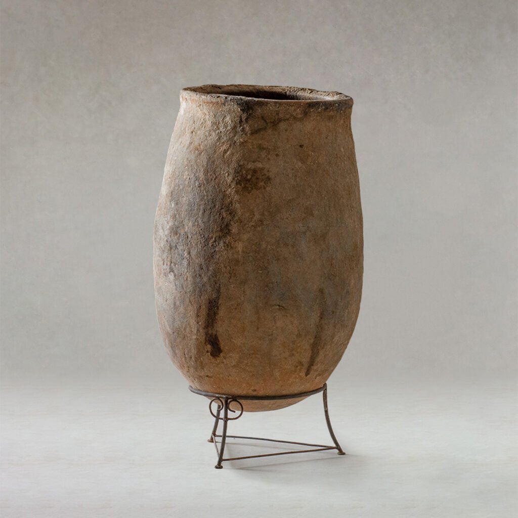 An ancient, tall, cylindrical clay vessel with a rough texture sits elegantly on a simple three-legged metal stand against a calming backdrop.