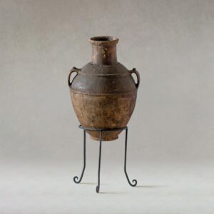 A rustic, two-handled clay urn with a narrow neck gracefully rests on a black wrought iron stand against a muted background. This timeless piece evokes a sense of artistry and tradition, adding an elegant touch to any setting.