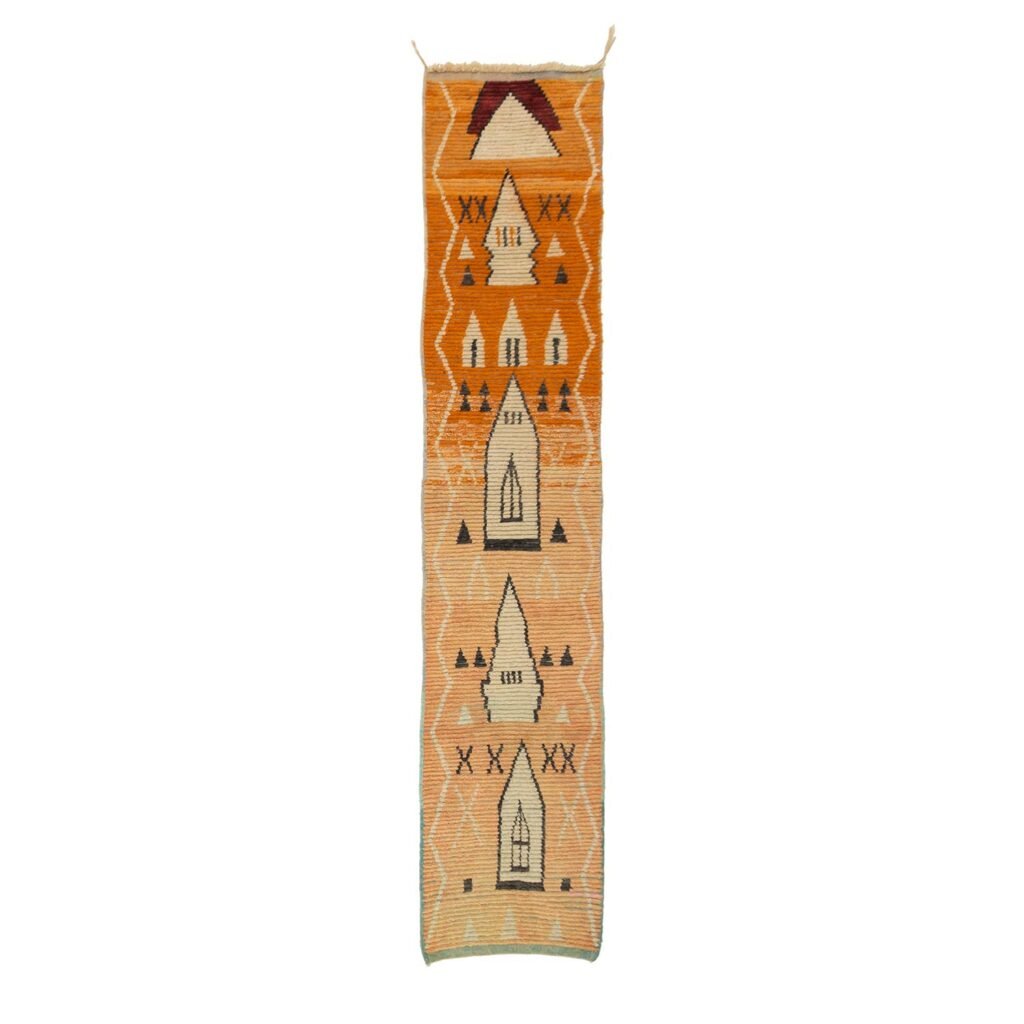 A long Berber runner rug showcasing geometric patterns in orange, tan, and brown tones. The design features triangles, diamonds, and abstract shapes.