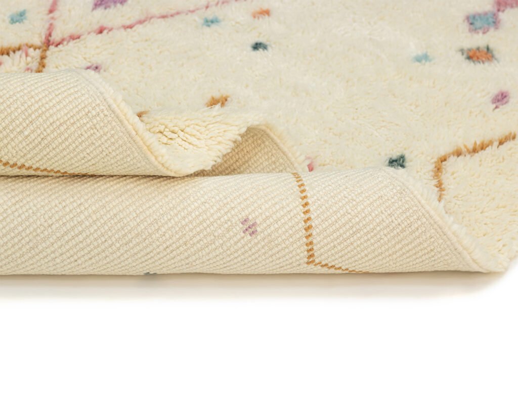 A folded corner of a luxury rug reveals its intricate underside, while the beige surface on top showcases colorful dots and accents.