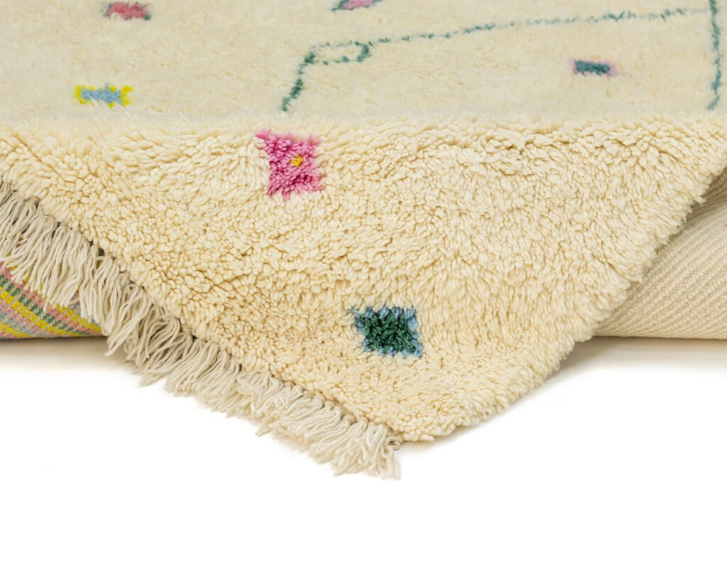 Close-up of a hand-knotted cream-colored rug with multicolored geometric patterns and fringed edges, partially folded over on a surface.